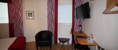 A compact hotel room with a bed, a desk, a chair, a TV, and a small table with a bottle.