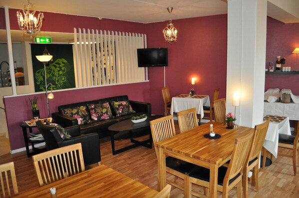 A dining area with a wooden table and chairs, a sofa with floral cushions, a chandelier, and a television mounted on the wall.