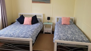 A room with two beds, each with patterned bedding and pillows, a small bedside table with a lamp, a painting on the wall, and a window with curtains. - Blue Haven - Pet friendly Cottage  (Iluka)