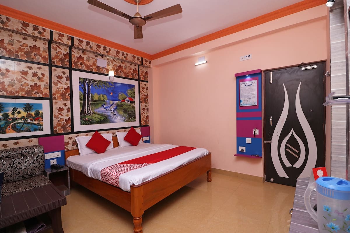 Habitación de hotel con cama, escritorio, silla, ventilador de techo, pared con cuadros y puerta con un cartel, además de un cubo de basura.