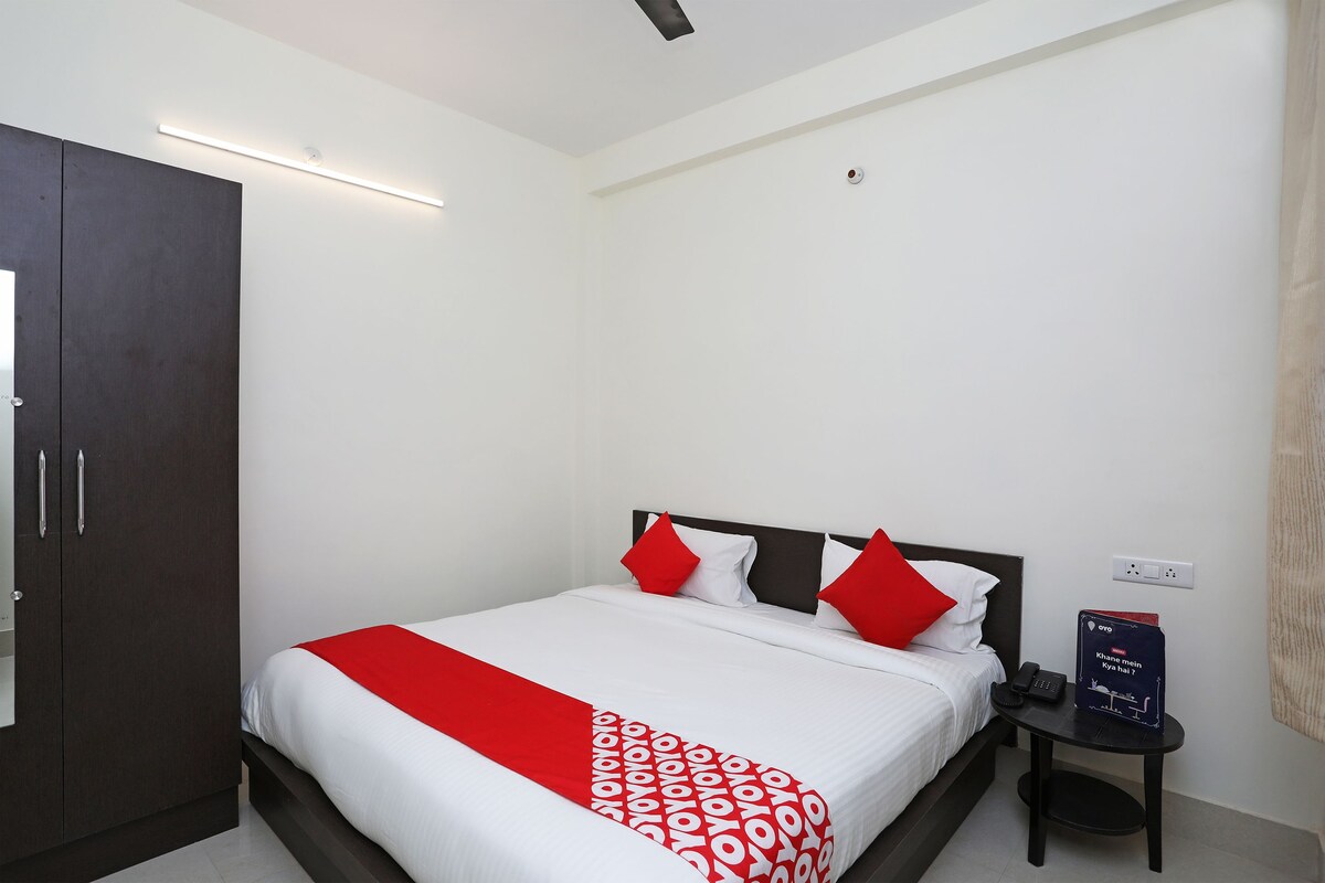 A neatly made bed with a red and white bedspread, a dark headboard, and red pillows. There is a small bedside table with a phone and a sign.