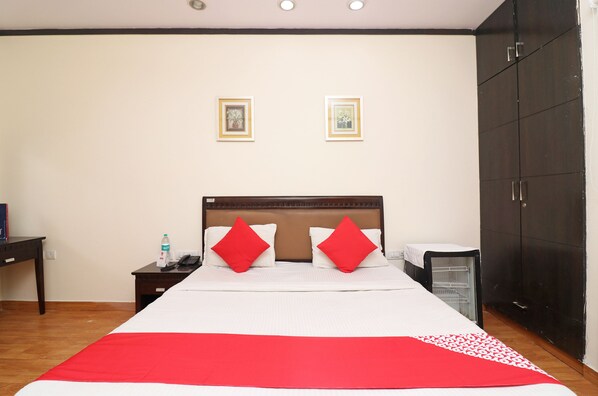 A neatly arranged hotel room with a large bed, two red pillows, a bedside table with a phone and water bottle, and a wardrobe.