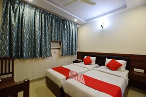Une chambre d’hôtel avec deux lits, une tête de lit en bois, une couvre-lit rouge et blanche, un ventilateur de plafond et une fenêtre avec des rideaux bleus.