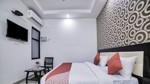 A hotel room with a bed, a TV, an air conditioner, a desk with a chair, and a patterned wall.