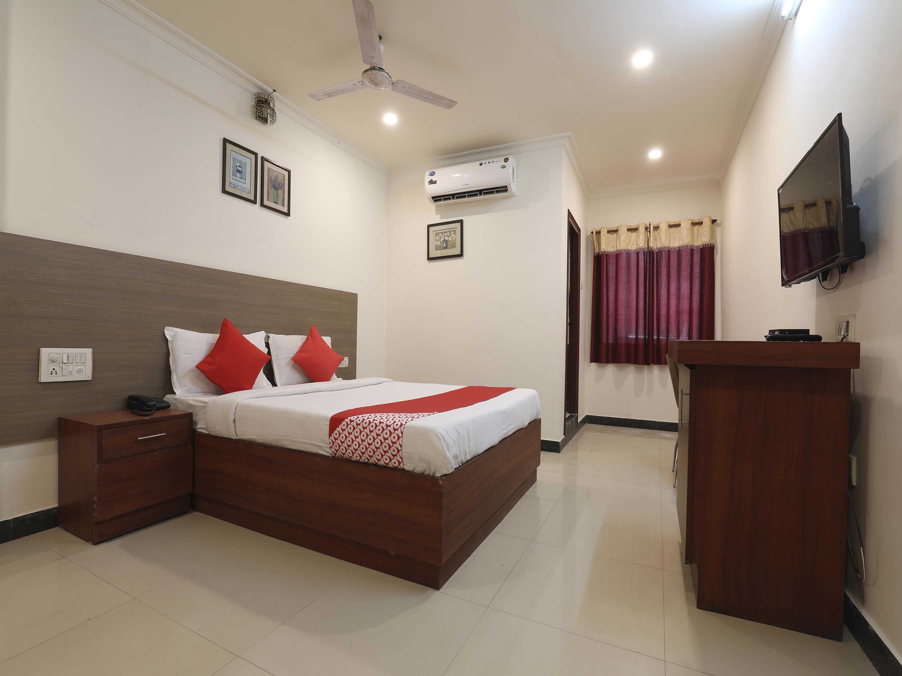 Une chambre d’hôtel comprenant un lit, une table de chevet, une commode, un climatiseur, un ventilateur de plafond, une télévision et un bureau en bois.