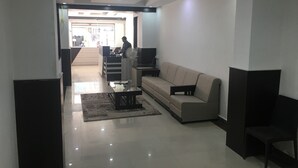 A modern living room with a sofa, a coffee table, and a TV. There is a person working at a desk in the background. - OYO 19909 Sri Guru Inn (Coimbatore)