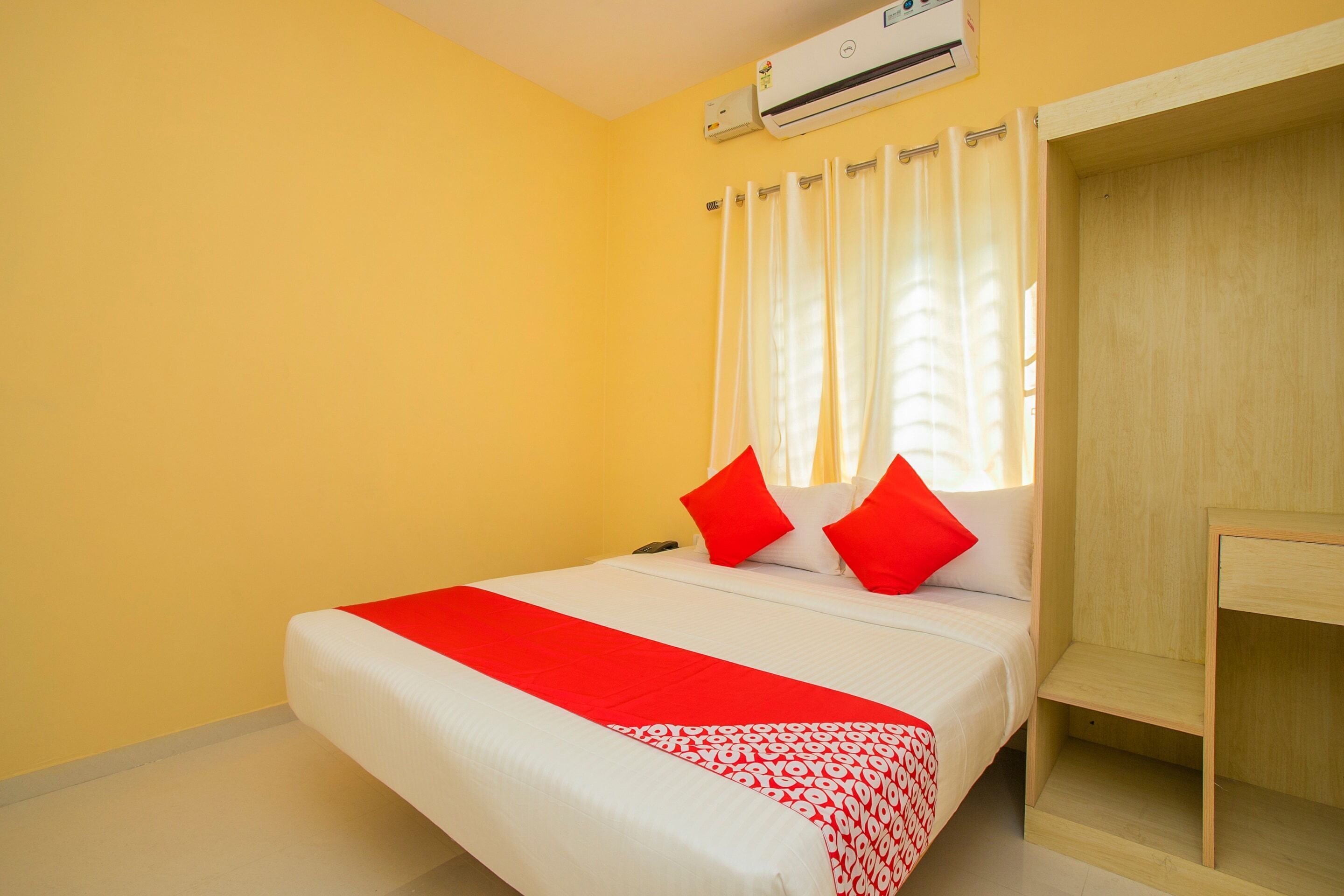 A hotel room with a bed, a bedside table, an air conditioning unit, and a window with curtains.