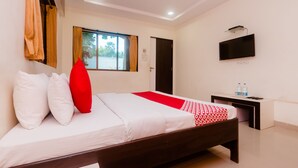 A neatly made bed with white and red bedding, a small bedside table with a TV and bottles, and a window with curtains. - OYO 12693 Manori Resort (Gorai)