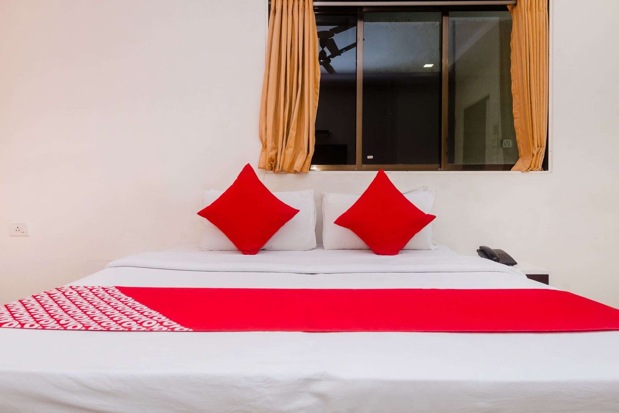 A bed with white bedding and two red pillows, a red blanket, and a window with curtains.