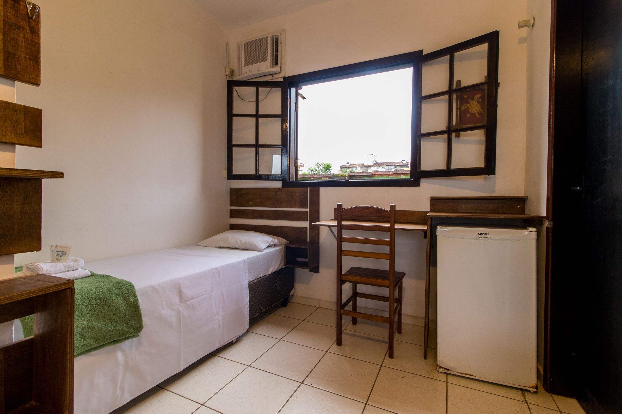 Una habitación pequeña y funcional con una cama individual, un escritorio, una silla y un refrigerador.