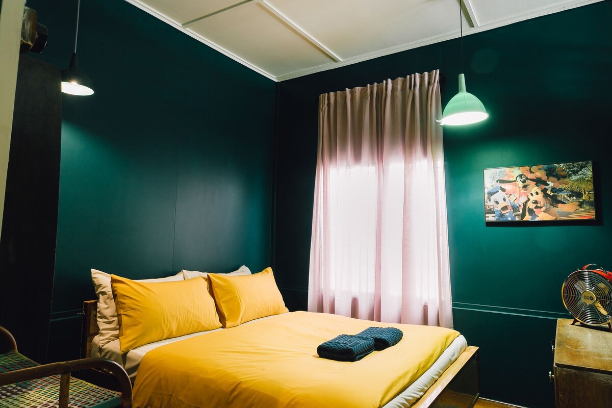 A room with a bed, a chair, a fan, and a painting on the wall.