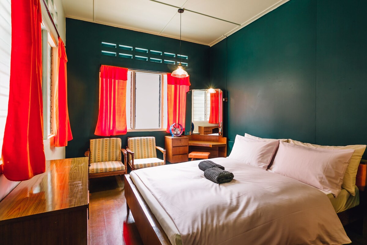 A hotel room with a bed, a desk, a chair, a dresser, and a window with red curtains.