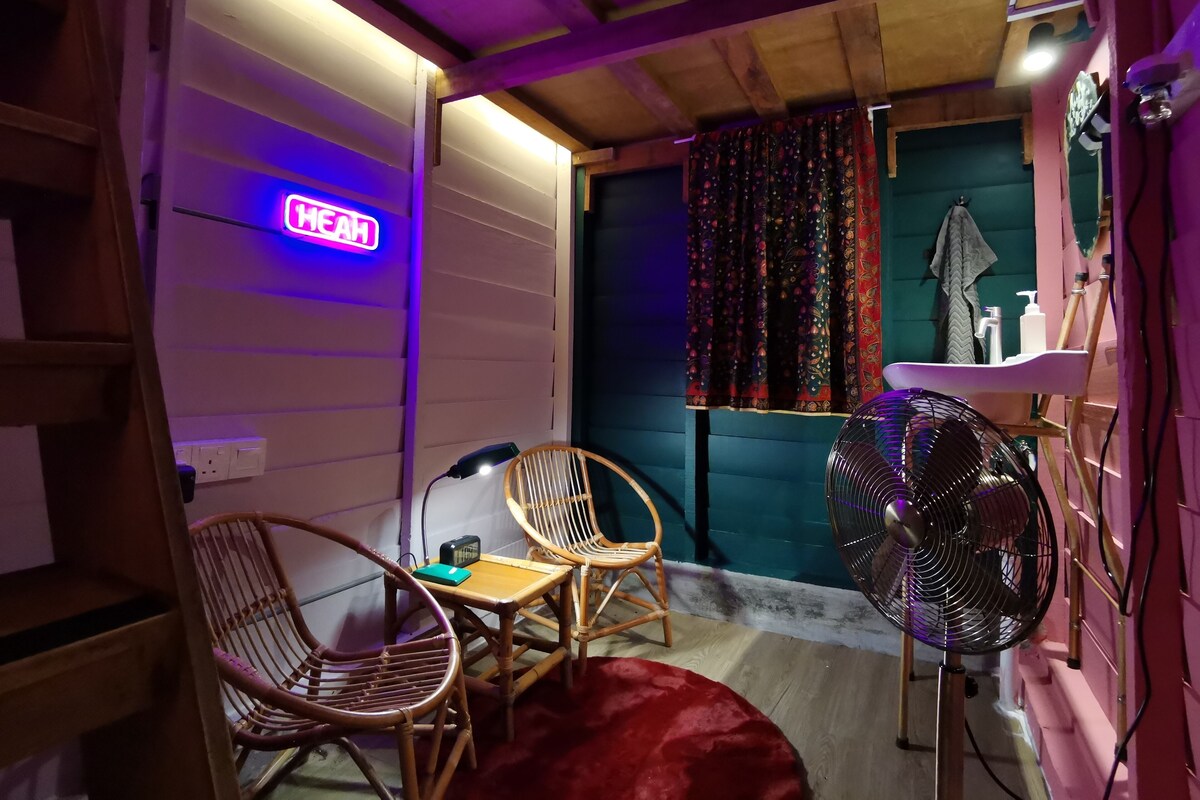 A cozy room with a neon sign, two wicker chairs, a small table, a red rug, and a standing fan.