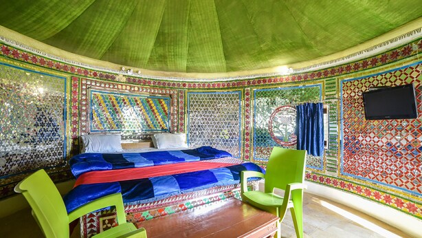 A room with a bed, two green chairs, a wooden table, and a TV on the wall. - OYO 24424 Kf Resorts Pvt Ltd (Jaisalmer)