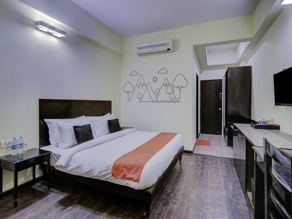 A hotel room with a bed, bedside tables, a desk, a TV, an air conditioning unit, and a wall with a mountain and tree mural.