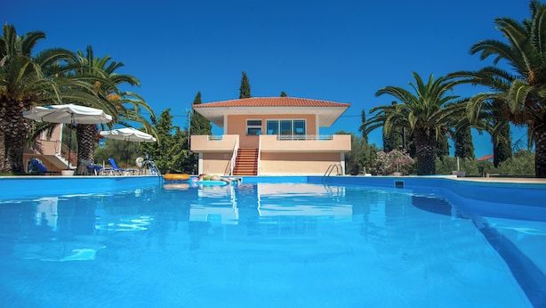 A swimming pool with a building in the background, surrounded by palm trees. - Helena Inn (Kefalonia)