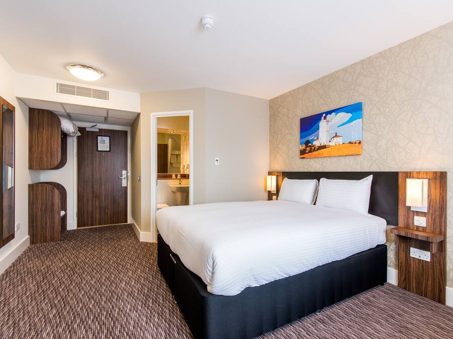 A hotel room with a large bed, a wooden headboard, a painting on the wall, and a carpeted floor.