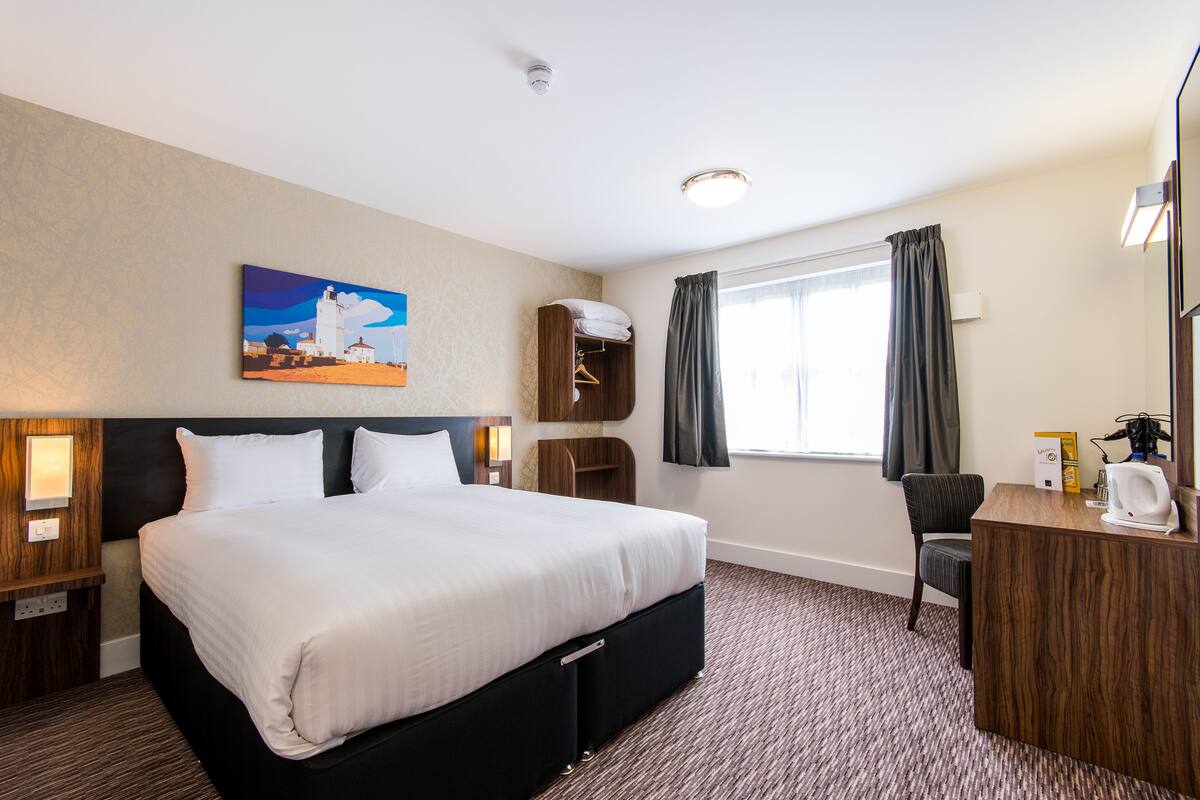 A hotel room with a large bed, a desk with a chair, a painting on the wall, and a window with curtains.