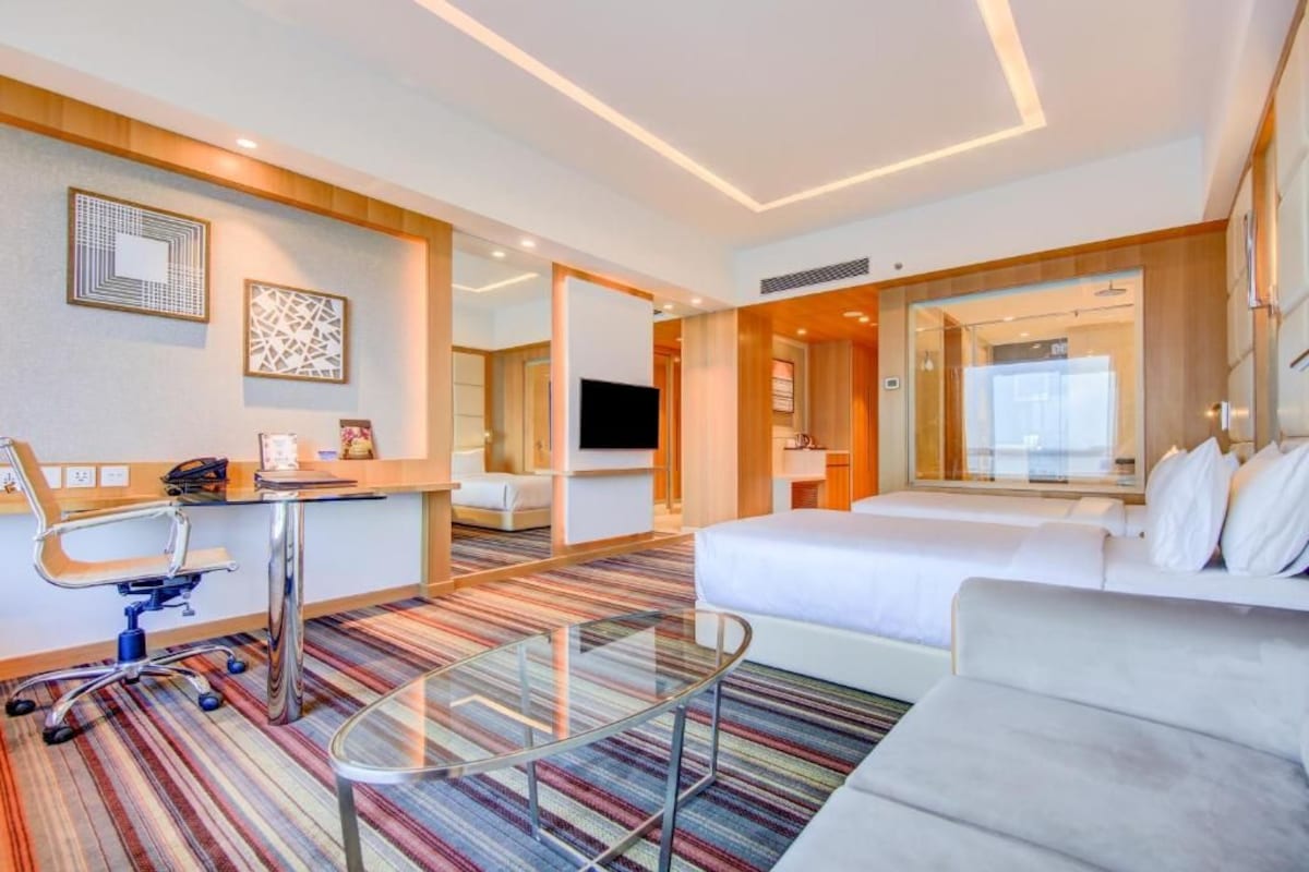 A hotel room with a bed, a sofa, a desk with a chair, a television, and a glass coffee table.