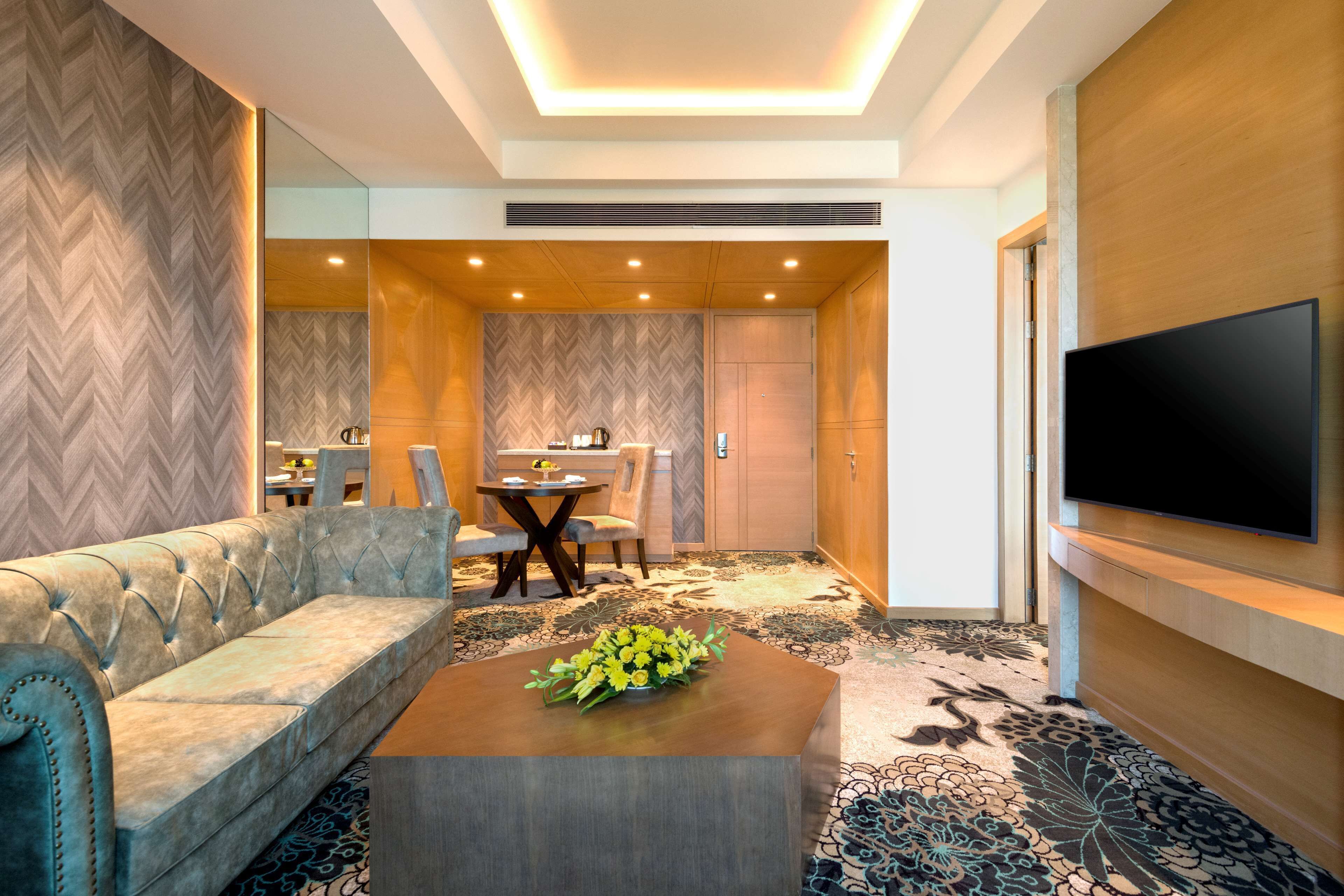 A modern hotel room with a sofa, a dining table with chairs, a flat-screen TV mounted on the wall, and a decorative flower arrangement on a coffee table.