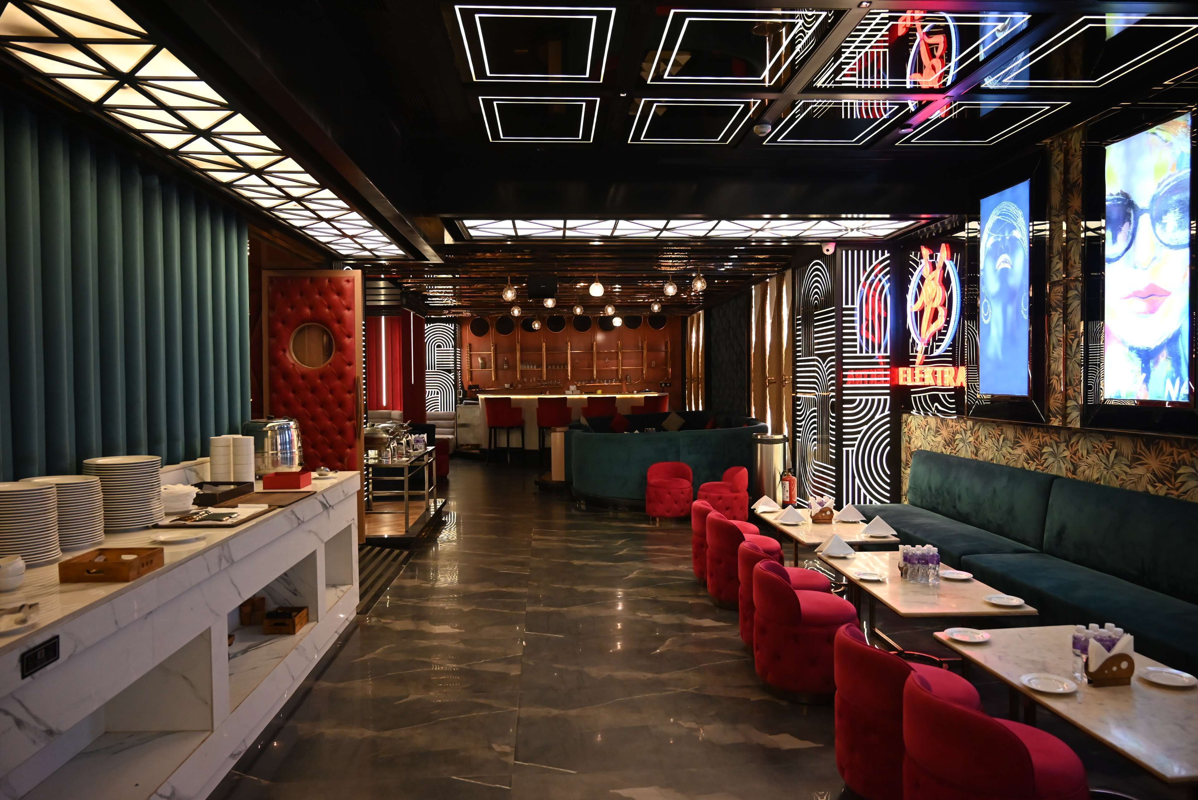 A restaurant with a modern interior, featuring a long counter with stacked plates and glasses, red upholstered chairs, and a green booth area.
