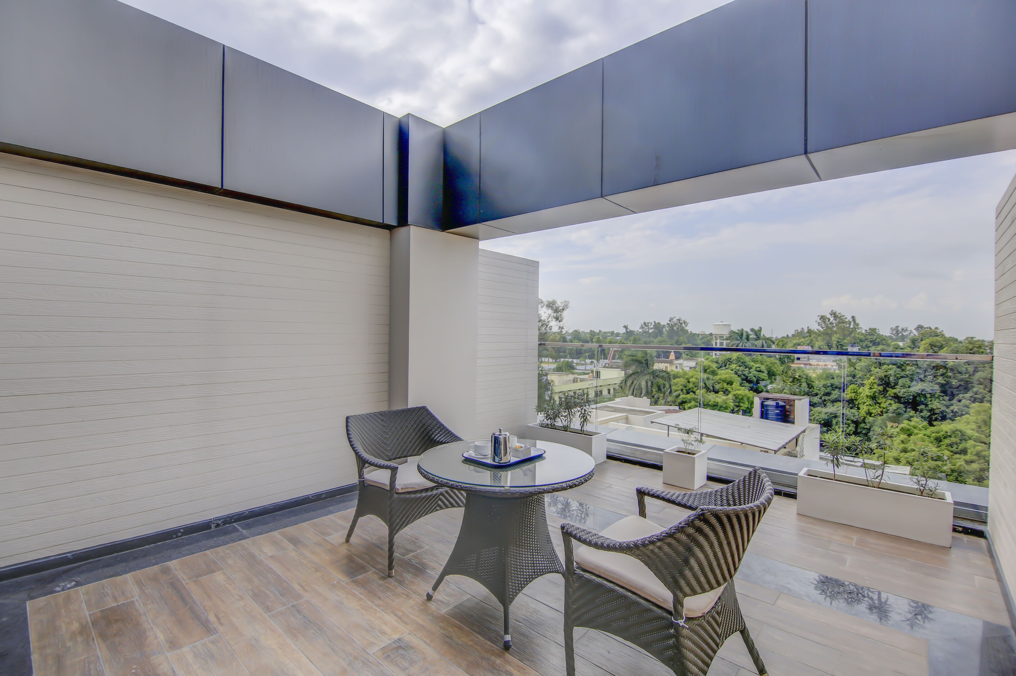Un balcón con mobiliario de mimbre, una mesa de vidrio y vistas a zonas verdes y edificios.