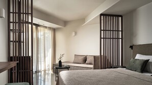 A modern hotel room with a large bed, a sofa, a small table with a vase, and a sliding door with vertical blinds. - Admiral De Luxe Hotel (Zakynthos)