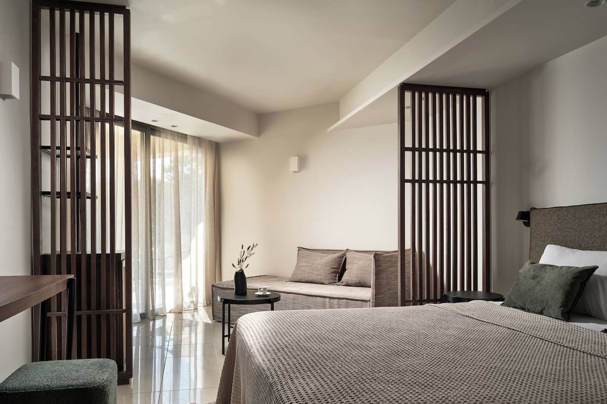 A modern hotel room with a large bed, a sofa, a small table with a vase, and a sliding door with vertical blinds.