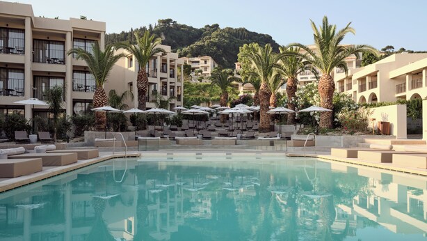 A pool area with lounge chairs and umbrellas, surrounded by palm trees and a building with balconies. - Admiral De Luxe Hotel (Zakynthos)