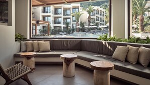A modern hotel room with a large window overlooking a pool and outdoor area, a comfortable seating area with a sofa and round wooden tables, and a view of a multi-story building with balconies. - Admiral De Luxe Hotel (Zakynthos)