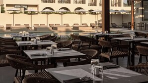 An outdoor dining area with wicker chairs and tables, a pool, and a building with balconies in the background. - Admiral De Luxe Hotel (Zakynthos)