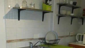 A compact kitchen with a sink, a small dining table, and a chair. There are shelves above the counter holding dishes and a green container. - Chocolate, March (Martos)