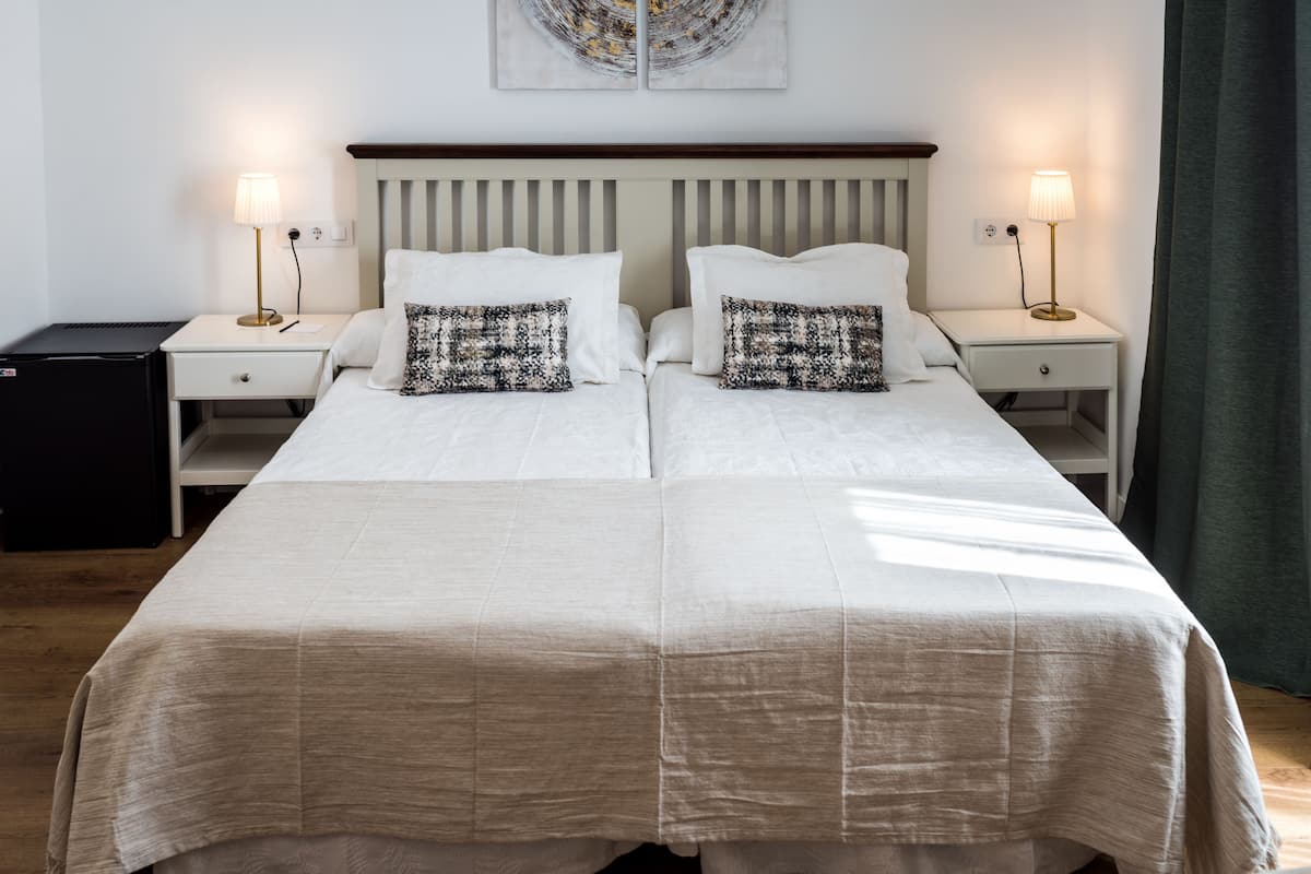 Een tweepersoonsbed met een beige sprei, twee nachtkastjes met lampen, een kleine zwarte koelkast en een aan de muur hangend kunstwerk.