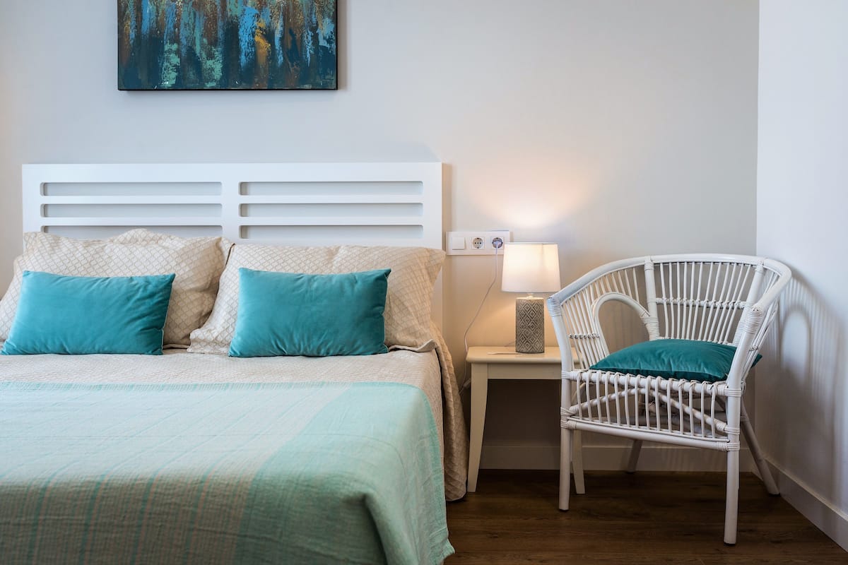 Een keurig opgemaakt slaapkamer met een bed, een witte nachtkastje met een lamp, en een witte rieten stoel.