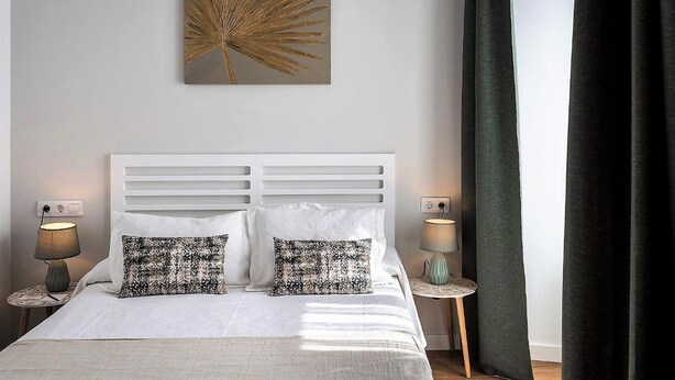 Een netjes opgemaakt bed met witte lakens en twee gestreepte kussens, met aan weerszijden nachtkastjes met lampen, in een kamer met een groot kunstwerk aan de muur en een donker gordijn.