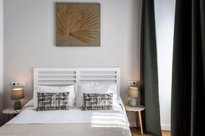 A neatly made bed with white linens and two patterned pillows, flanked by two bedside tables with lamps, in a room with a large wall art piece and a dark curtain.