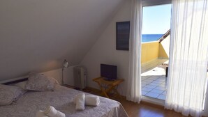 Une chambre à coucher avec un lit, des oreillers, une télévision et une vue sur la mer, visible par la porte-fenêtre donnant sur le balcon.