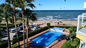 Une piscine entourée de palmiers, une plage animée où l’on voit des gens et des cerfs-volants, et une vue sur l’océan.