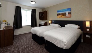 A hotel room with two beds, a desk, a coat rack, and a painting of a lighthouse.