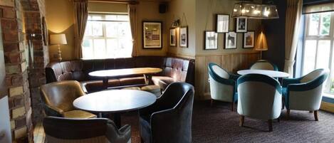 A cozy lounge area with leather sofas, round tables, and chairs, featuring a brick wall and framed pictures on the walls.