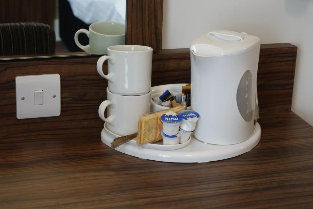 A coffee maker, a stack of white cups, and a tray with food items on a wooden surface.