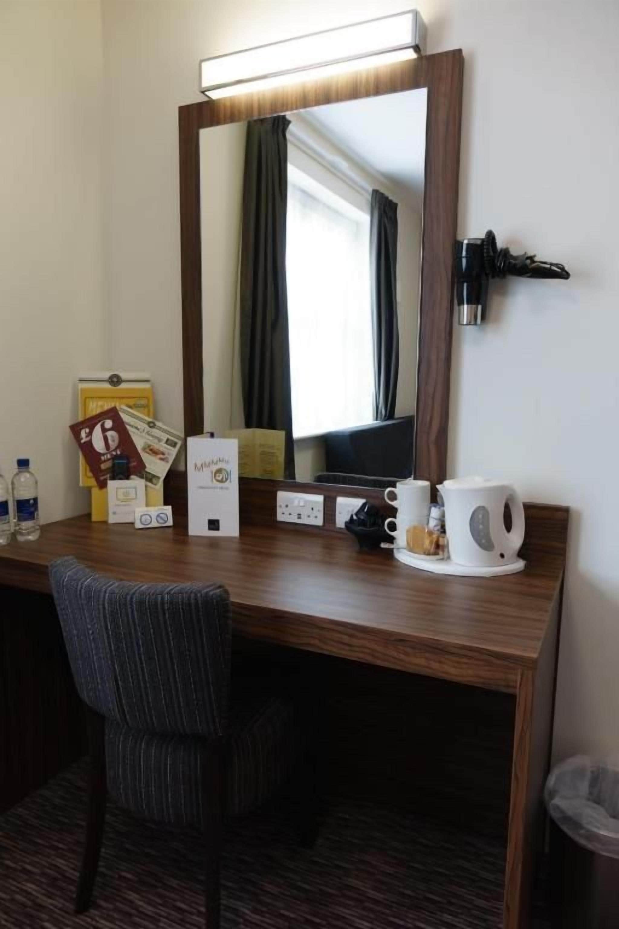Une chambre d’hôtel avec un bureau en bois, une chaise, un miroir, une machine à café, une poubelle et une fenêtre avec des rideaux.
