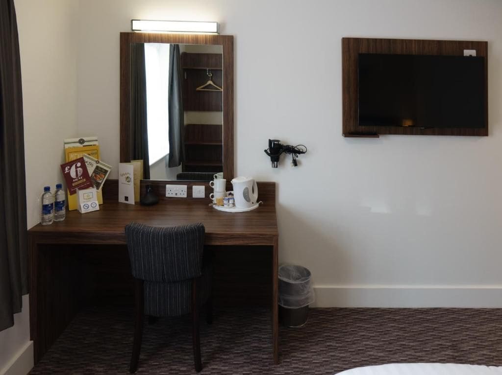 A hotel room with a wooden desk, a chair, a mirror, a television, and a small trash bin.