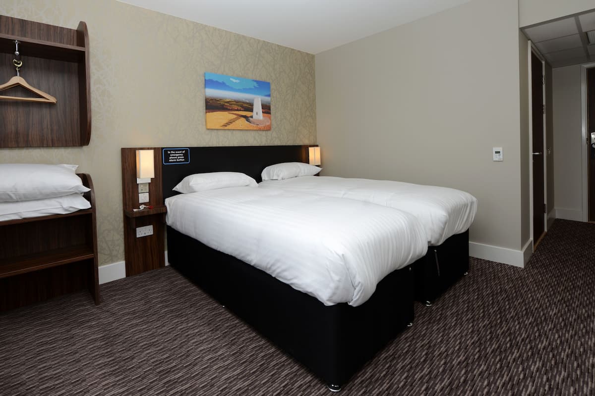 Une chambre d’hôtel avec un grand lit, une commode en bois, une table de chevet, une lampe fixée au mur, un tableau et une porte.