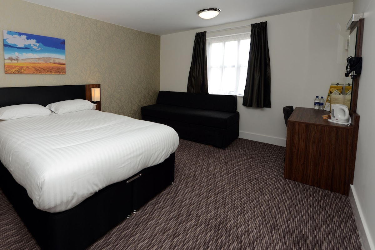 A hotel room with a large bed, a sofa, a small desk with a kettle, and a painting on the wall.