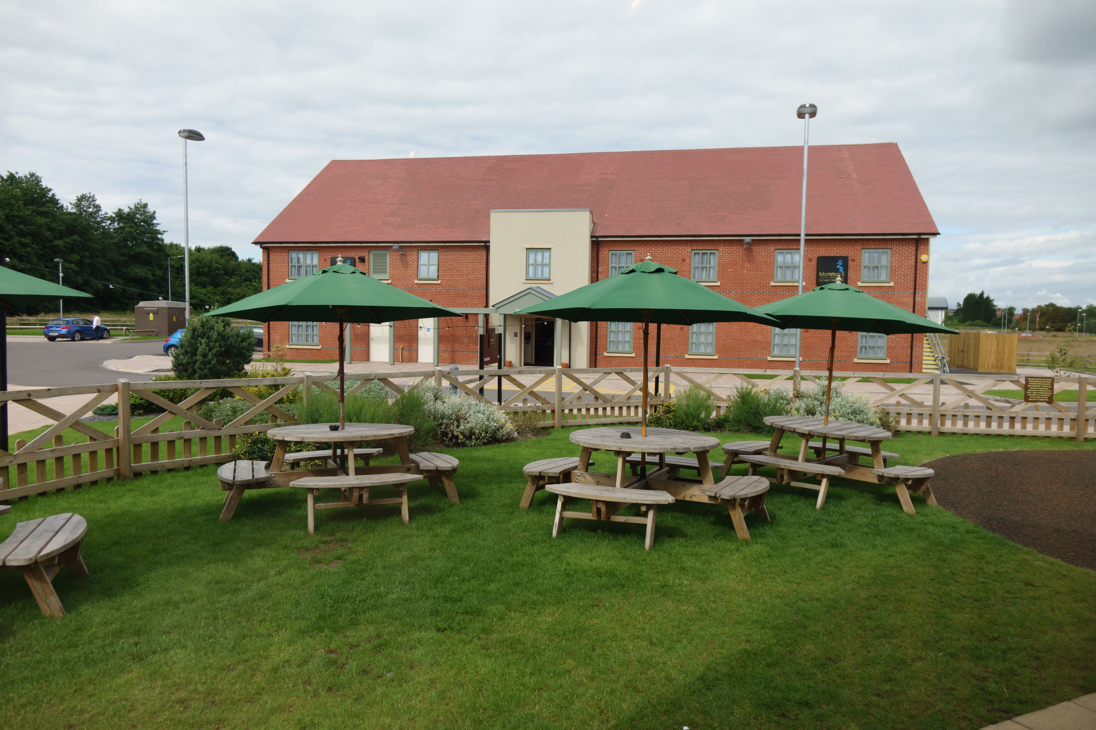 A building with a red roof and a brick facade, surrounded by a wooden fence and a grassy area with picnic tables and green umbrellas.