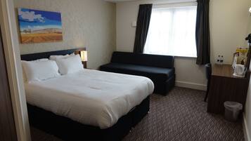 A hotel room with a large bed, a sofa, a desk with a lamp, a trash can, and a window with curtains.