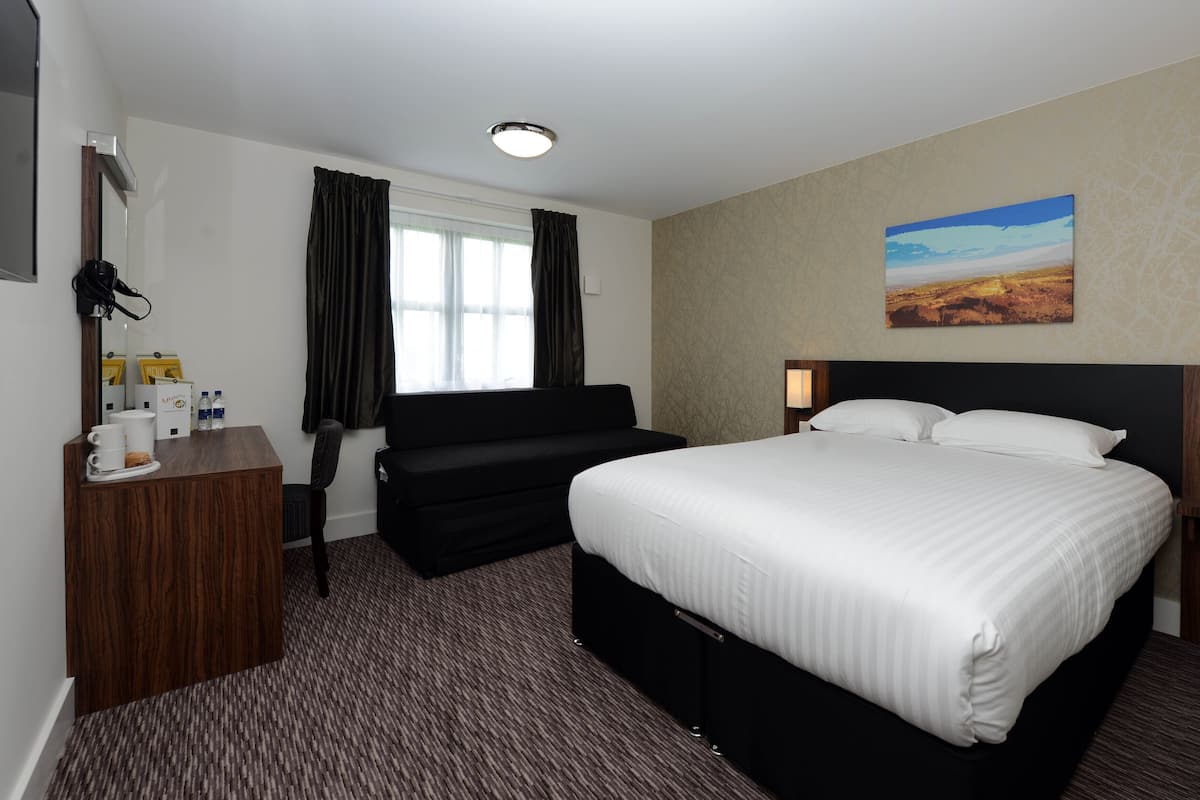 Une chambre d’hôtel avec un grand lit, un bureau avec une chaise, une fenêtre avec des rideaux, un tableau au mur et un sol recouvert de moquette.
