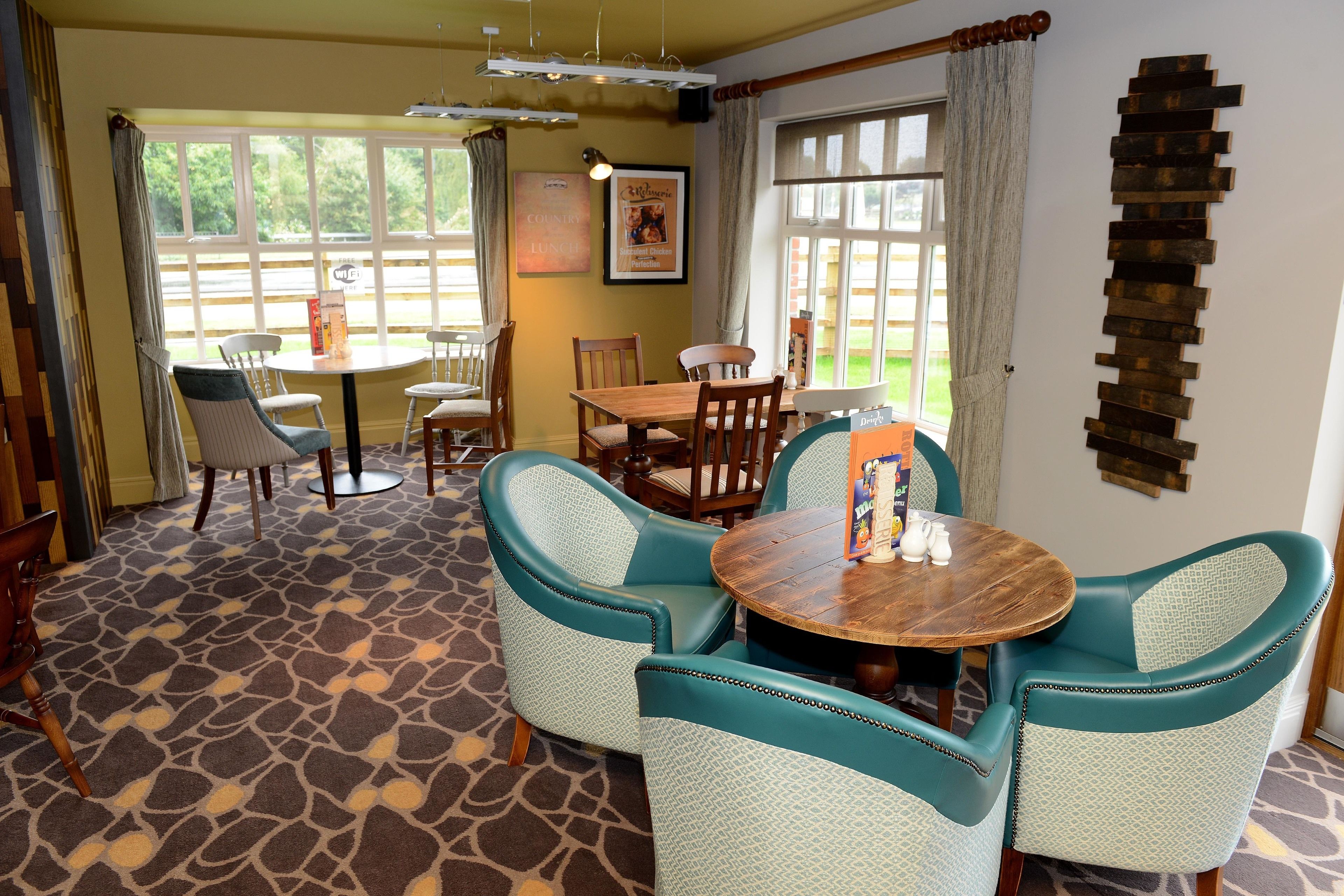 A dining area with a round wooden table surrounded by teal upholstered chairs, a patterned carpet, and a view of greenery through large windows.