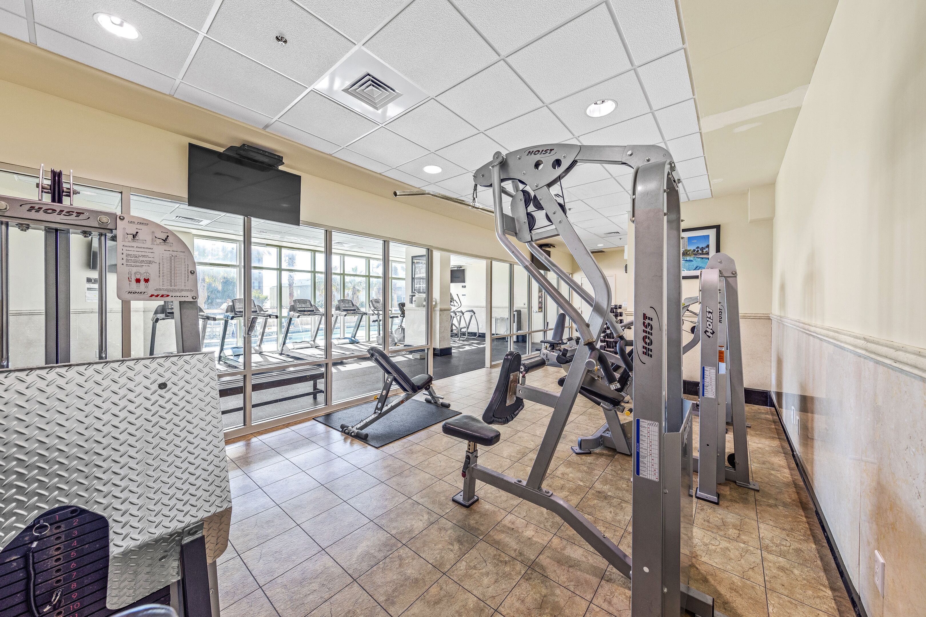 A gym with various exercise equipment, including weight machines and treadmills, with large windows allowing natural light.